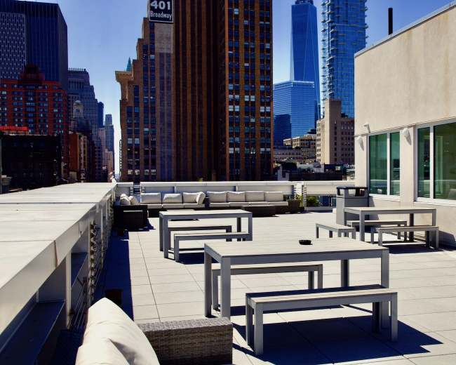 A rooftop terrace with gray tables, benches, and a clear view of tall buildings in the background.