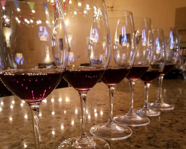 A row of wine glasses filled with red wine sits on a granite countertop under colorful party decorations.