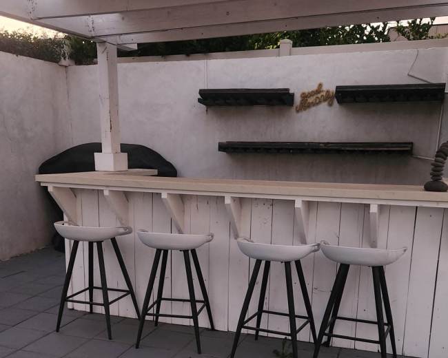 The image shows a patio area with a white bar counter featuring four white stools and a bare wall in the background with shelves.