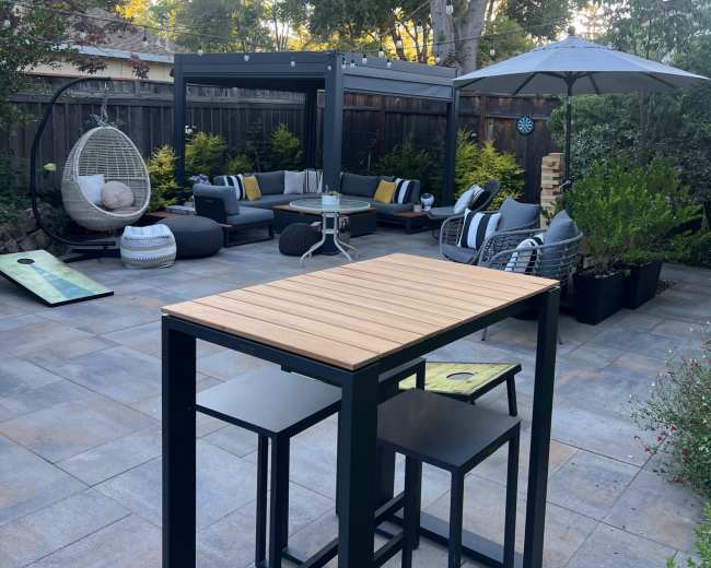 The image shows a spacious outdoor patio area with modern seating arrangements, a bar table, and greenery surrounding the space.