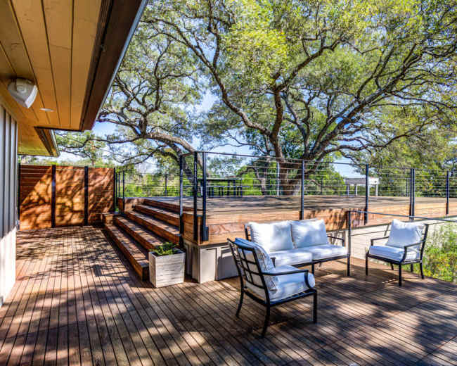 A wooden deck with seating overlooks a landscaped area and large trees under bright blue skies.