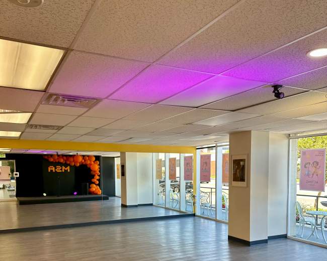 The image shows a spacious, well-lit room with a wooden floor, large windows, and colorful decorations, including orange balloons and poster displays.