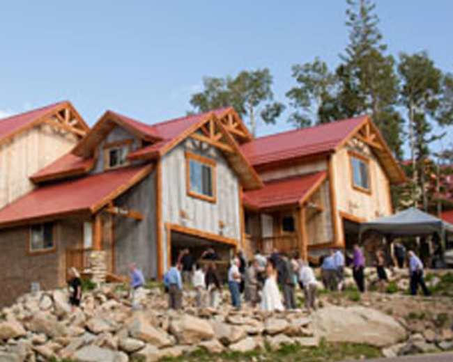 A rustic lodge with wooden structures and a red metal roof is surrounded by trees and hosts a gathering of people outside.