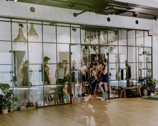 A group of people is engaged in a fitness class inside a modern studio with large glass walls and wooden flooring, surrounded by indoor plants.
