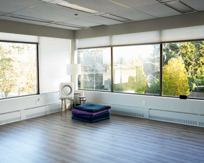 A bright, empty room with large windows, hardwood floors, a lamp, and cushions arranged on the floor.