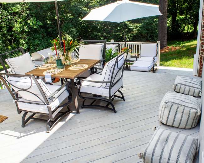 A wooden patio features a dining table with chairs, an umbrella, and additional seating on a deck surrounded by trees.