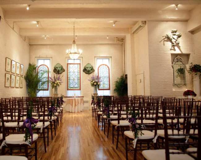 The image shows a decorated indoor venue set up for a wedding ceremony, featuring rows of wooden chairs facing a beautifully adorned altar with floral arrangements and stained glass windows.