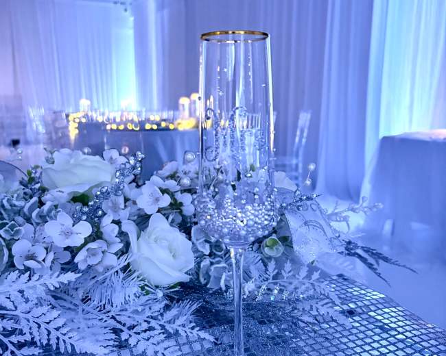 A decorated champagne flute stands on a shimmering table setting adorned with white roses and silver foliage under ambient blue lighting.