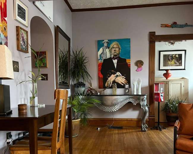 A wooden table and chair are positioned in a cozy living room, featuring a large painting on the wall and various plants throughout the space.