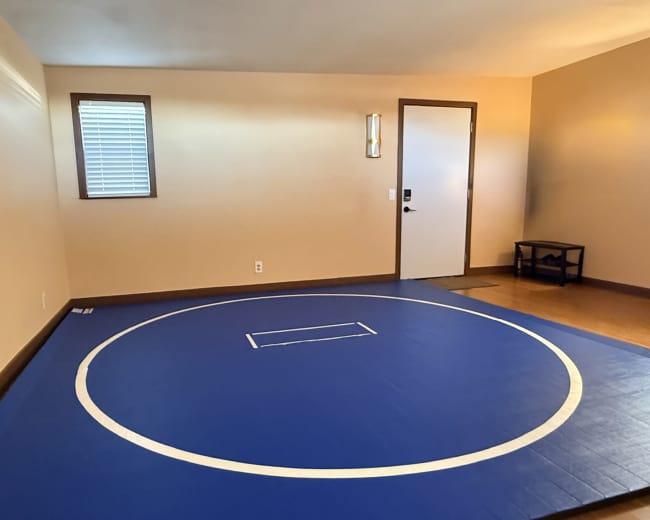 The image shows a room with wooden flooring that features a blue wrestling mat with a white circle and a small rectangle in the center.