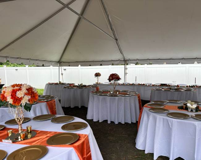 The image shows a tented event space with round tables covered in white tablecloths, adorned with orange and gold decorations and plates.