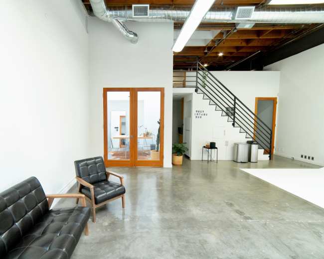 The image shows a modern interior space with a black leather sofa, a wooden armchair, a staircase, and large glass doors leading to another room.