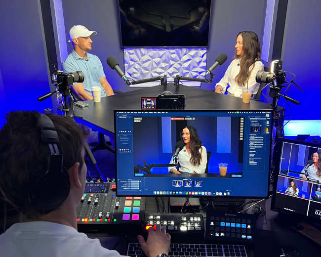 A man and a woman are seated at a table during a podcast recording, while a third person operates a camera and controls the audio equipment in a well-lit studio.