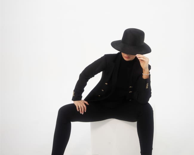 A woman in a black suit and hat sits on a white cube, with her legs crossed and one hand adjusting her hat.