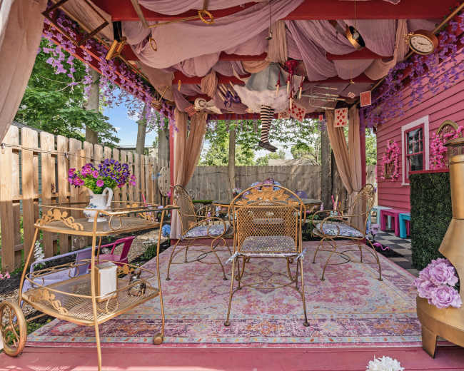 The image shows a decorated outdoor gazebo with draped fabric, flowers, and gold furniture arranged around a patterned rug.