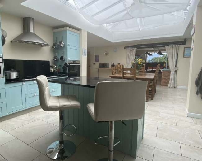 The image shows a modern kitchen featuring light blue cabinetry, a black countertop, and a seating area with two white bar stools, all illuminated by natural light from a skylight.