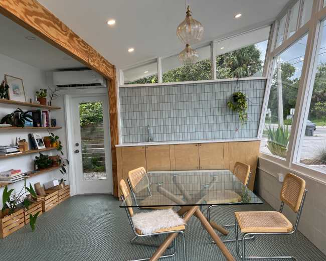 A modern kitchen dining area features a glass table surrounded by four cane chairs, with a wall of windows providing natural light.