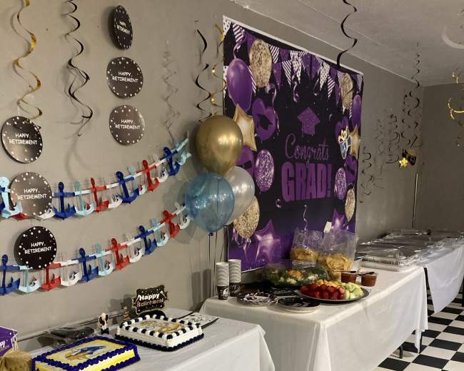 The scene shows a decorated party space with tables covered in white cloths, displaying a variety of foods, drinks, and a festive backdrop celebrating a graduation.