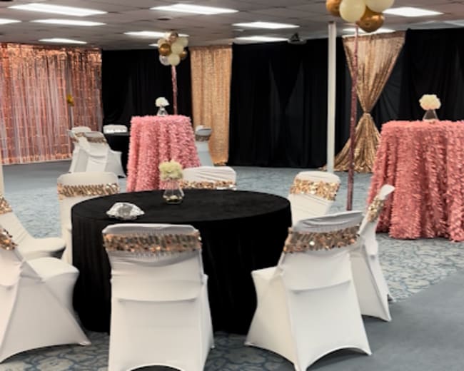 The image shows a decorated event space with tables covered in pink and black tablecloths, surrounded by white chairs.