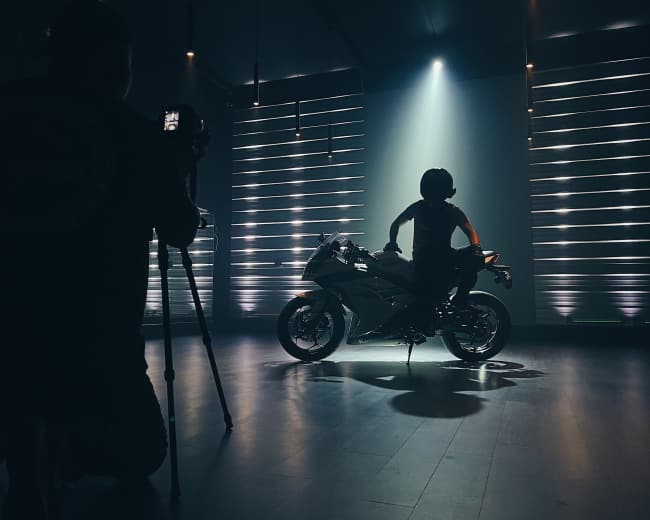 A person is photographing a motorcyclist posing on a motorcycle under dramatic stage lighting.