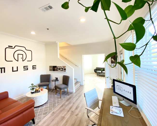 The image shows a modern office space with a large wall logo that reads "MUSE," featuring a seating area, a desk with a computer, and a staircase in the background.