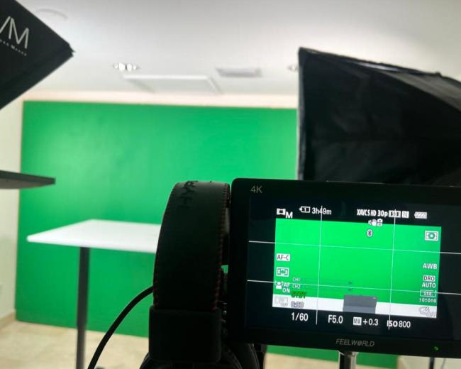 A camera setup with a monitor displaying a live view of a green screen and a table, with lighting equipment positioned on either side.