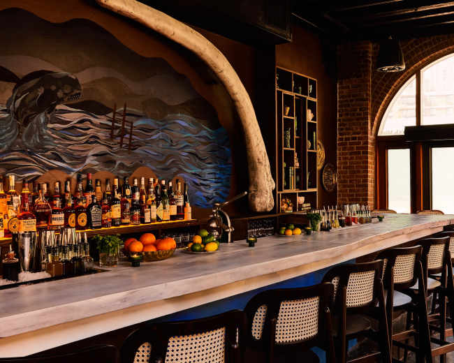 The image shows a bar with a long marble countertop, several bottles of liquor neatly arranged on shelves, and a large mural featuring waves and a whale skull.