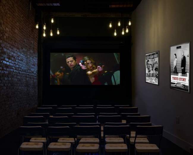 A small movie theater features empty rows of folding chairs facing a large screen displaying a scene from a film, with vintage movie posters on the walls.