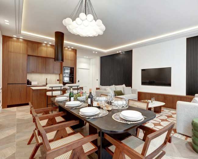 The image shows a modern dining area with a large table set for a meal, surrounded by wooden chairs, and a living space featuring a sofa and television in a contemporary design.