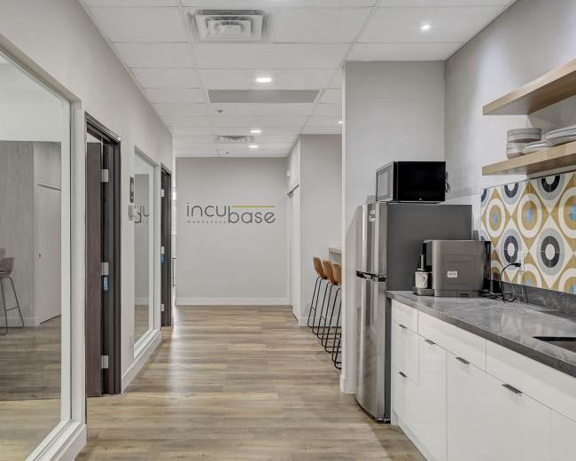 A modern office hallway leading to a kitchenette and several glass-walled offices, with a logo reading "incubase" on the wall.