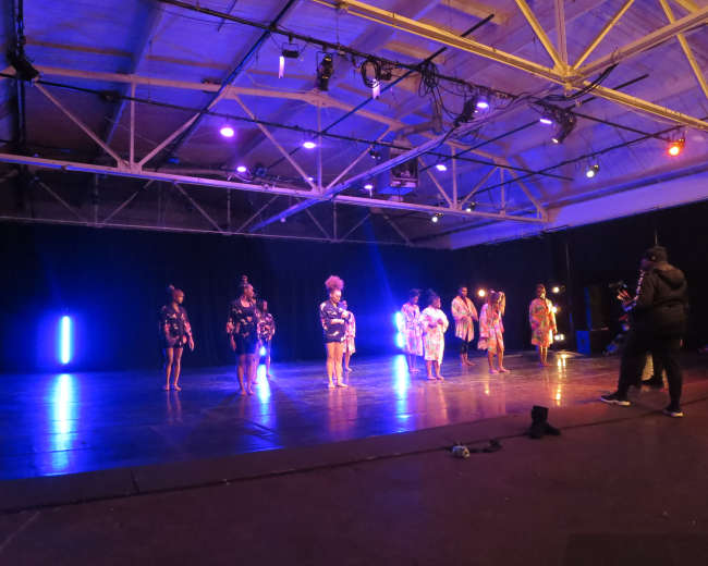 A group of performers dressed in costumes stands on a stage with various lighting effects during a dance rehearsal.