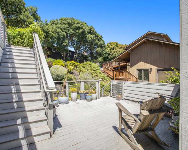 A wooden deck with stairs leads to a landscaped backyard featuring potted plants and a neighboring house.