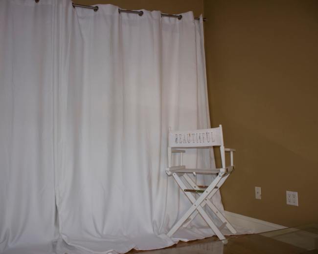 A white chair labeled "BEAUTIFUL" sits in front of a set of white curtains against a beige wall.