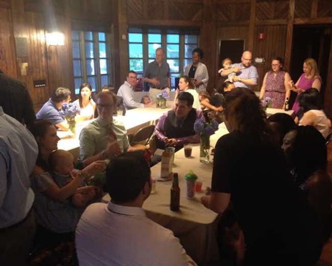 The image shows a group of people gathered around tables in a wooden room, with some holding babies and others engaged in conversations.