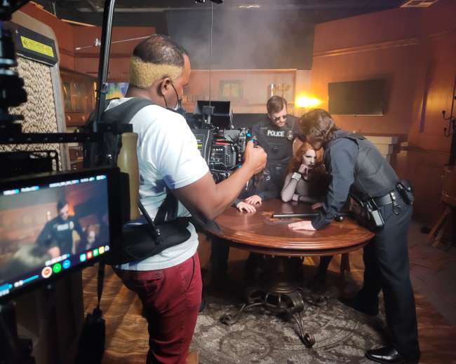 A film crew captures a scene with police officers and a suspect gathered around a table in a dimly lit room.