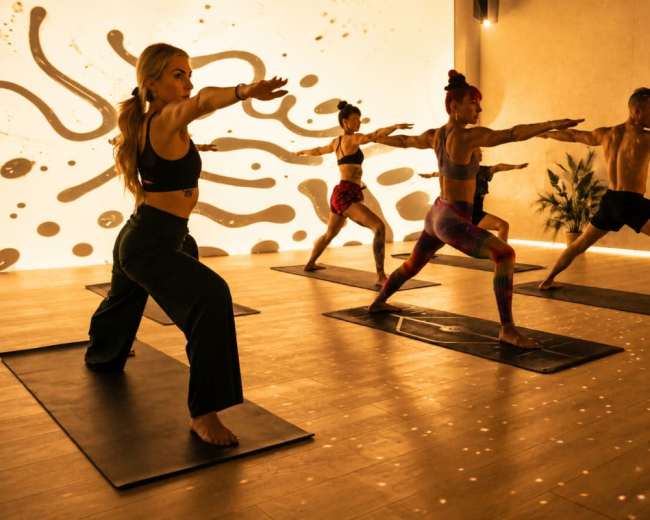 A group of individuals is practicing yoga in a brightly lit studio with abstract wall designs, each person in a different pose on their mats.