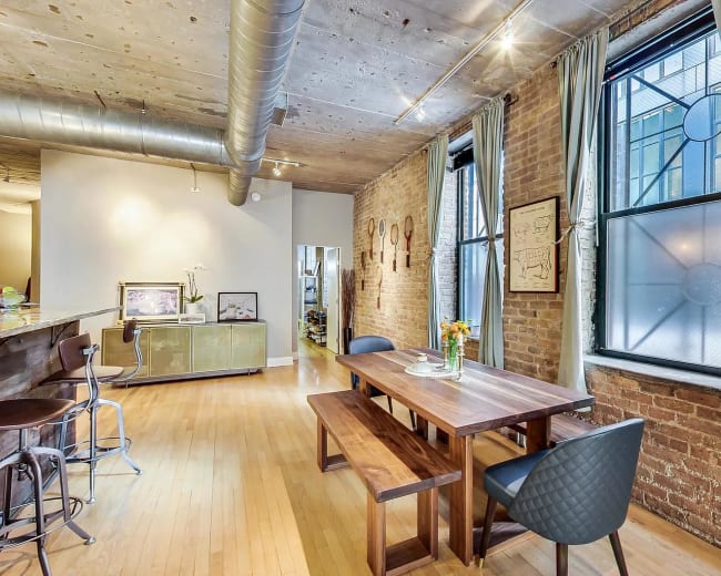 The image shows a modern dining area with a wooden table, chairs, and large windows featuring exposed brick walls.