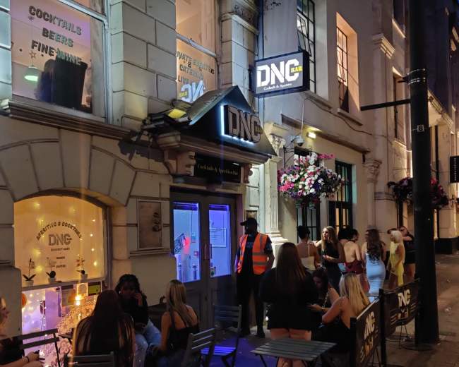 The image shows a bar entrance labeled "DNG" with patrons sitting outside on a street lined with flowers.