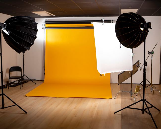 The image shows a photography studio with a yellow backdrop, two light setups, and a foldable chair.