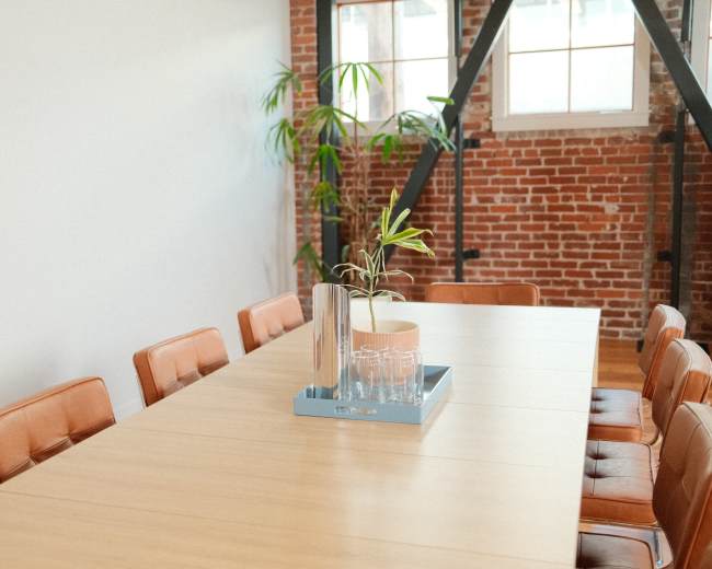 A long wooden conference table is surrounded by brown leather chairs in a room with exposed brick walls and natural light.