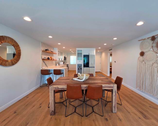The image depicts a modern dining area with a wooden table surrounded by six brown chairs, adjacent to an open kitchen featuring sleek cabinetry and a large window.