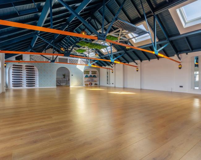 The image shows a spacious interior with a wooden floor, high ceiling beams painted blue, and large windows providing natural light.