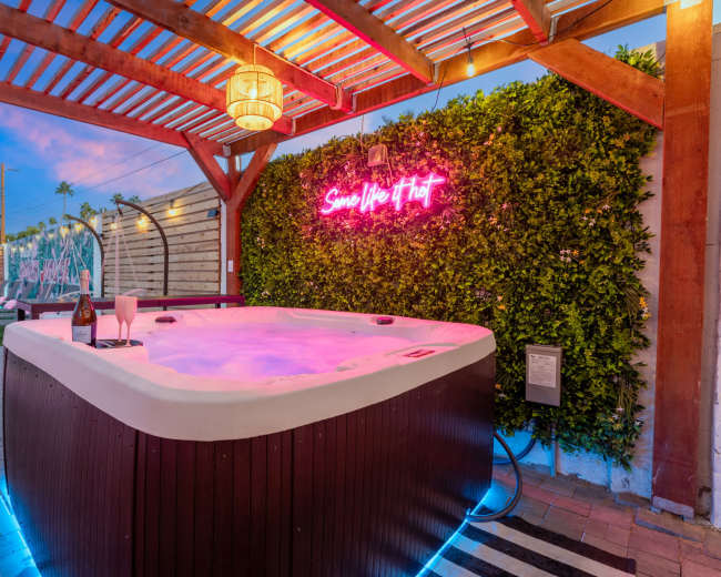A hot tub with pink lighting is situated under a wooden pergola, next to a green wall with a neon sign, and surrounded by lounge chairs and outdoor decor.