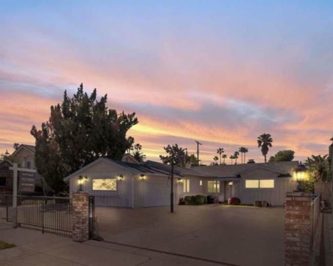 A modern single-story house is pictured with a driveway enclosed by a gate, set against a colorful sunset sky.