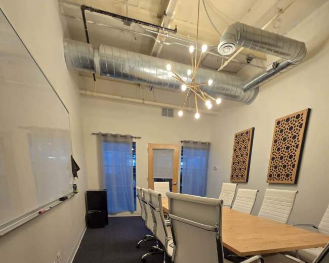 A conference room with a long wooden table surrounded by white chairs, featuring a whiteboard and artwork on the walls.