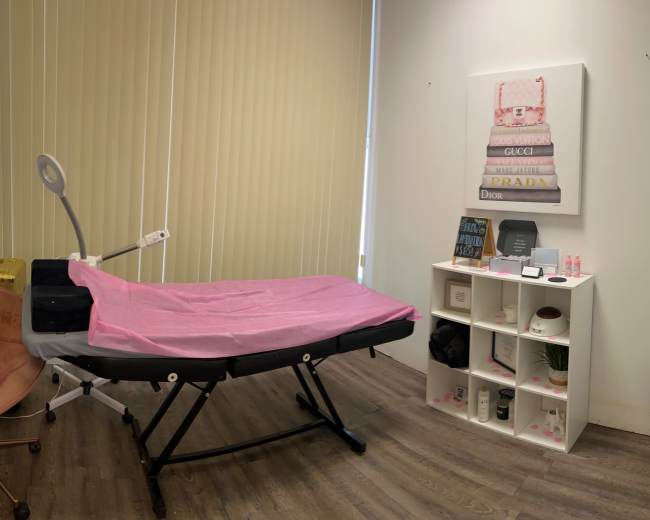 The image shows a treatment room with a massage table covered in a pink sheet, a chair, and a storage unit displaying various beauty products and decorations.