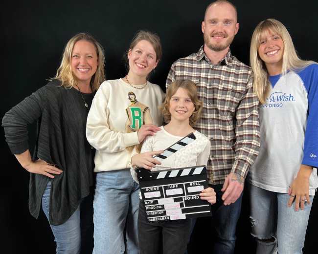 A group of five people, including a child holding a clapperboard, pose together in front of a black backdrop.