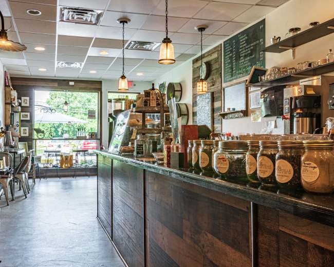 A coffee shop interior features a wooden counter, jars on display, and various seating arrangements, with large windows overlooking an outdoor area.