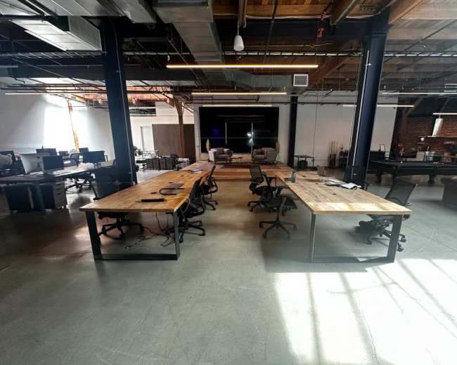 The image shows a modern open office space with large wooden tables, black office chairs, and a few empty workstations under high ceilings and natural light.