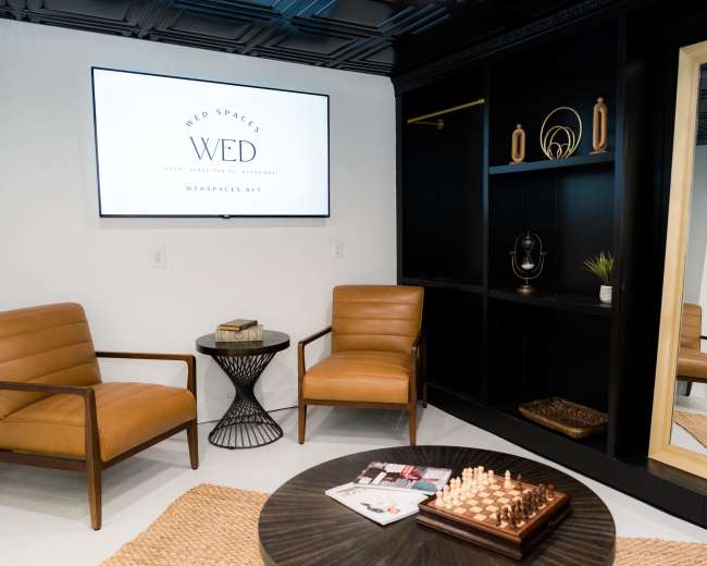 The image shows a modern waiting area with two brown chairs, a round table, and a large mirror, featuring an overhead television displaying the logo for "Web Spaces."